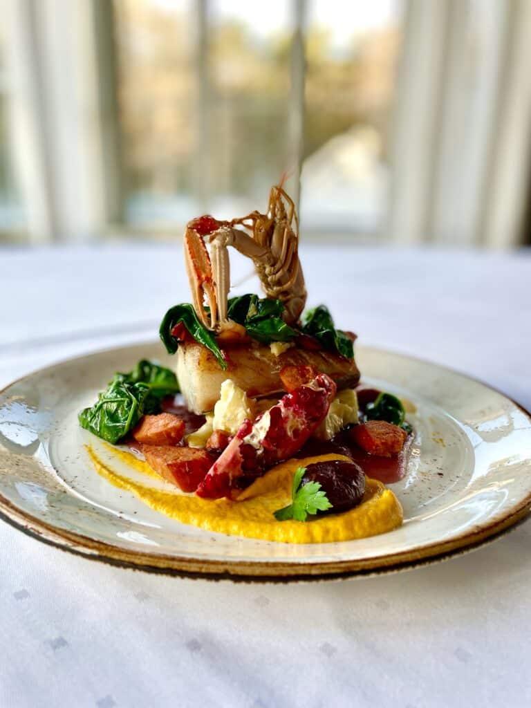 Gourmet sjømatrett med sjøkreps kamskjell og fargerike grønnsaker på en hvit tallerken med gullkant.