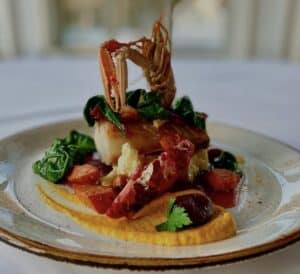 Gourmet sjømatrett med sjøkreps kamskjell og fargerike grønnsaker på en hvit tallerken med gullkant.